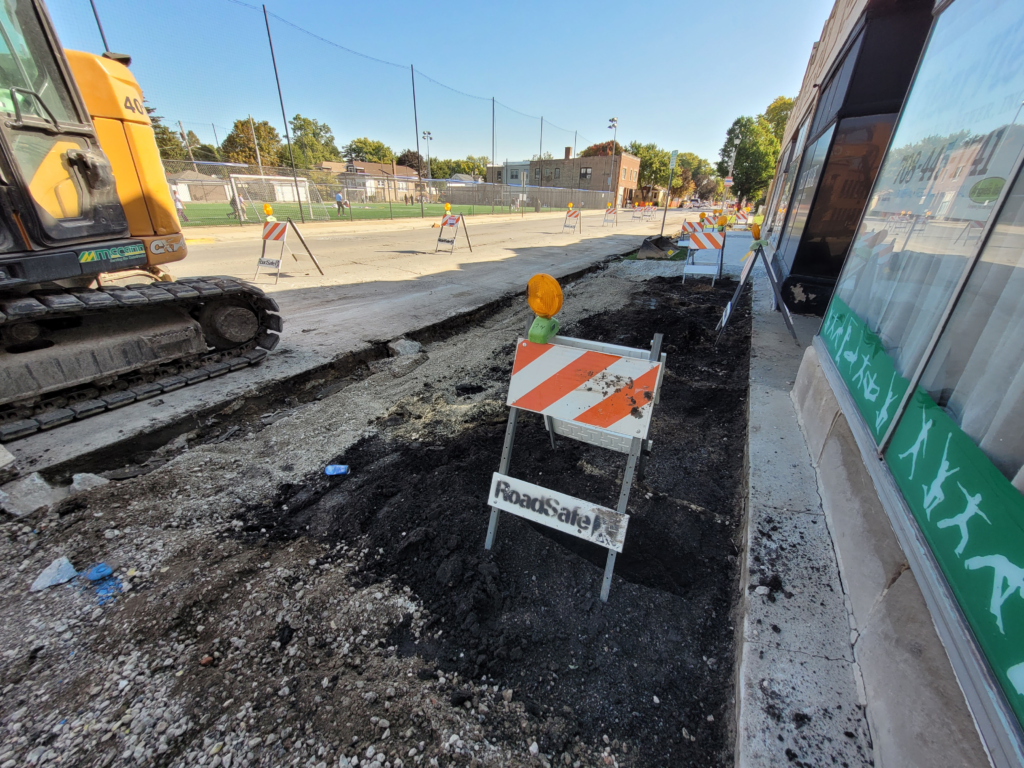 Excavated sidewalk area beside a building along 16th Street, with an excavator and construction barricade.