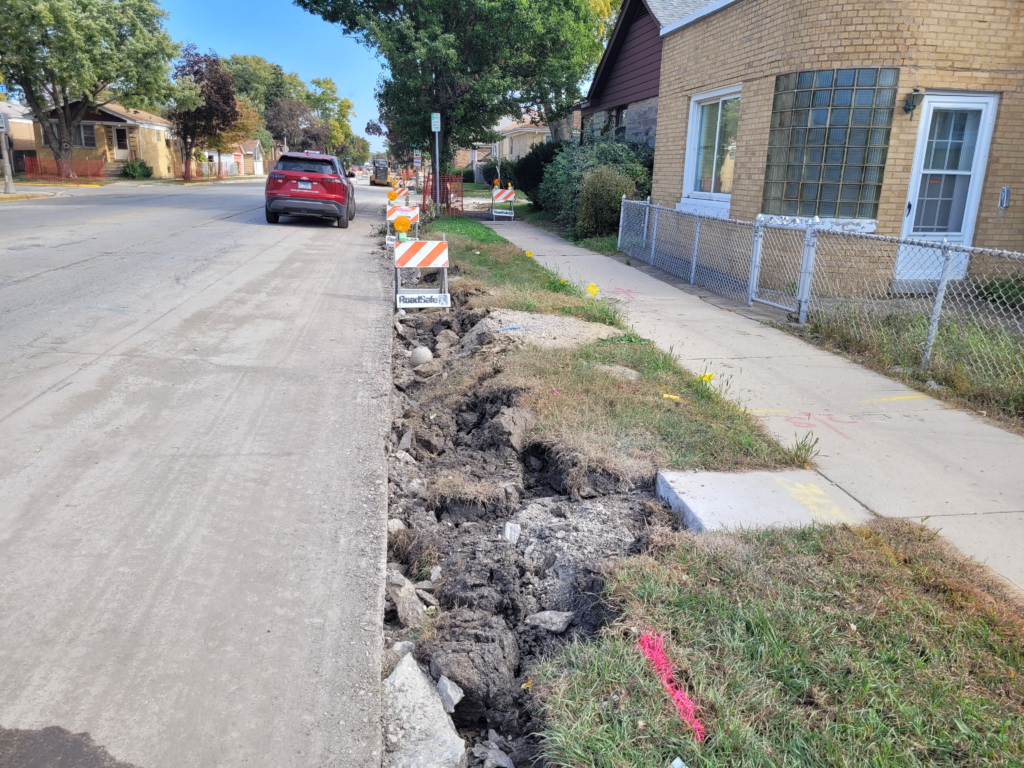 Excavated curb line along 16th Street following curb and gutter removal, with construction barricades beside the sidewalk.