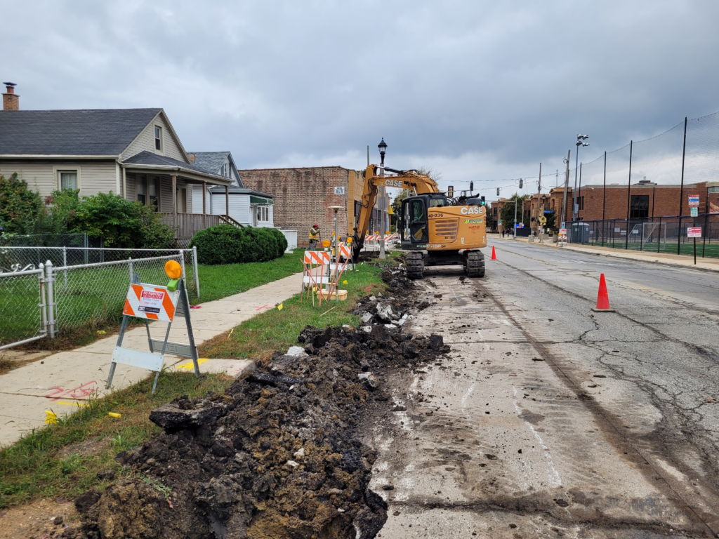 Excavator along 16th Street beside removed curb and gutter, with soil and broken pavement along the roadway edge.