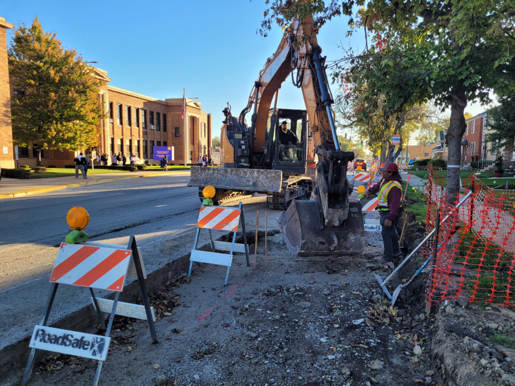 Excavator working in a removed sidewalk area along 16th Street, with barricades and tree protection fencing nearby.
